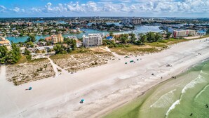 Vista aérea de una zona costera con una playa de arena extensa, un puerto deportivo con barcos y edificios a lo largo de la orilla.