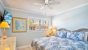 Une chambre à coucher avec un lit, deux lampes de chevet, un ventilateur de plafond, une fenêtre avec des stores et un tableau encadré au mur.