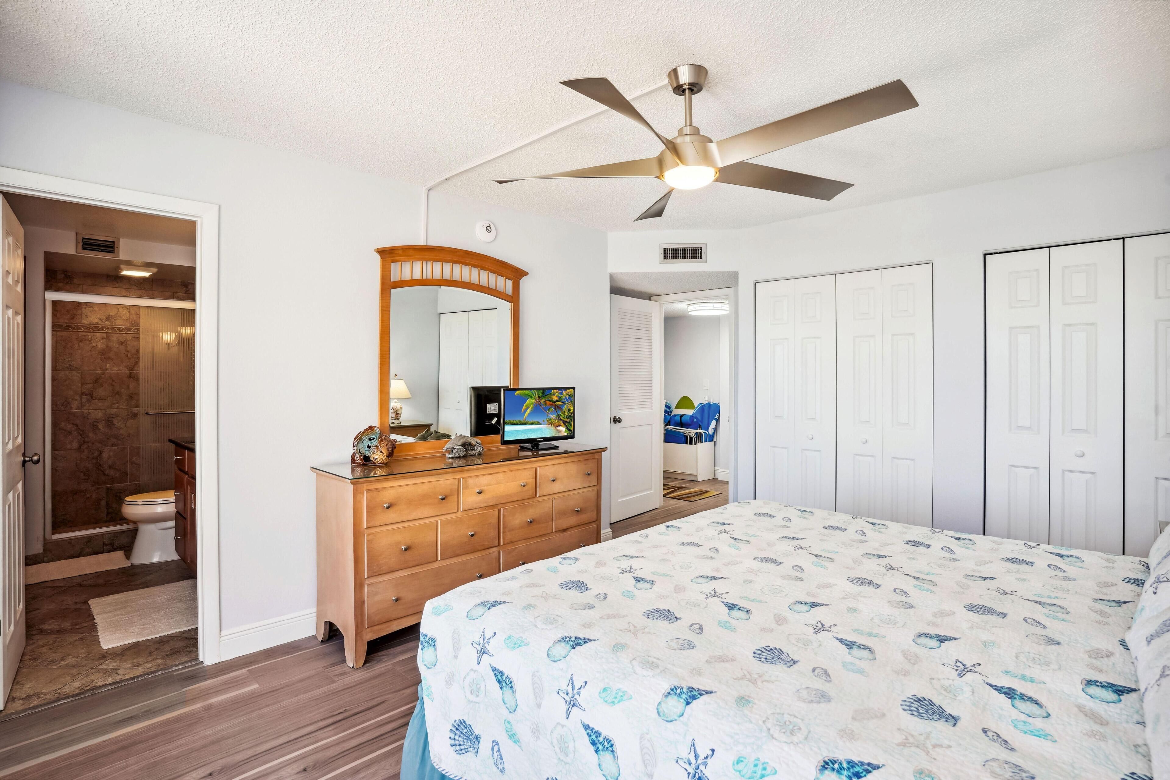 Ein Schlafzimmer mit Bett, Deckenventilator, Kommode, Fernseher und einem durchgehenden Türrahmen einsehbarer Badezimmer.