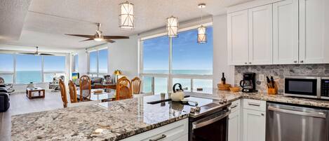A modern kitchen with granite countertops, stainless steel appliances, and a large window overlooking the ocean.