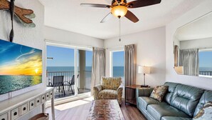 A spacious living room with a view of the ocean, a ceiling fan, a leather sofa, a patterned ottoman, and a wooden coffee table. - Sunset Vistas 603w (Treasure Island)