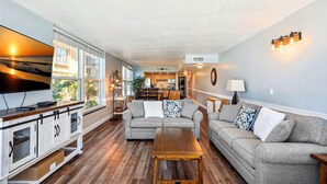 A spacious living room with a large window, a flat-screen TV, and a wooden coffee table. - Madeira Vista 201 (Madeira Beach)