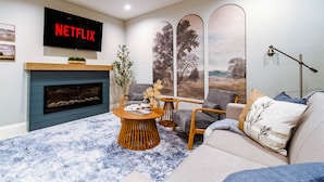 A living room with a fireplace, a television mounted on the wall, and a large window with a view of trees.