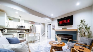 A modern living room with a kitchen, a fireplace, and a TV mounted on the wall displaying "NETFLIX".