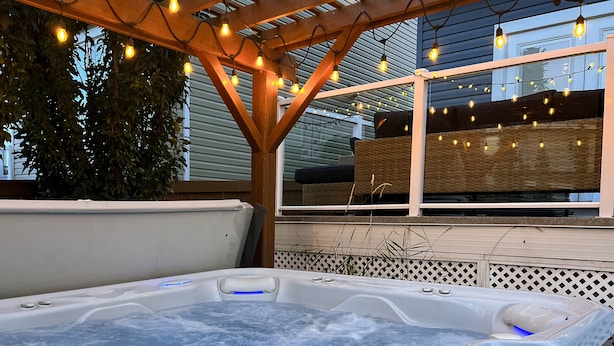 A hot tub under a wooden pergola with string lights, surrounded by a wooden deck and a view of a house.