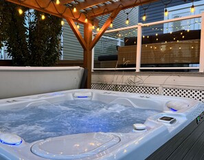 A hot tub under a wooden pergola with string lights, surrounded by a wooden deck and a view of a house.