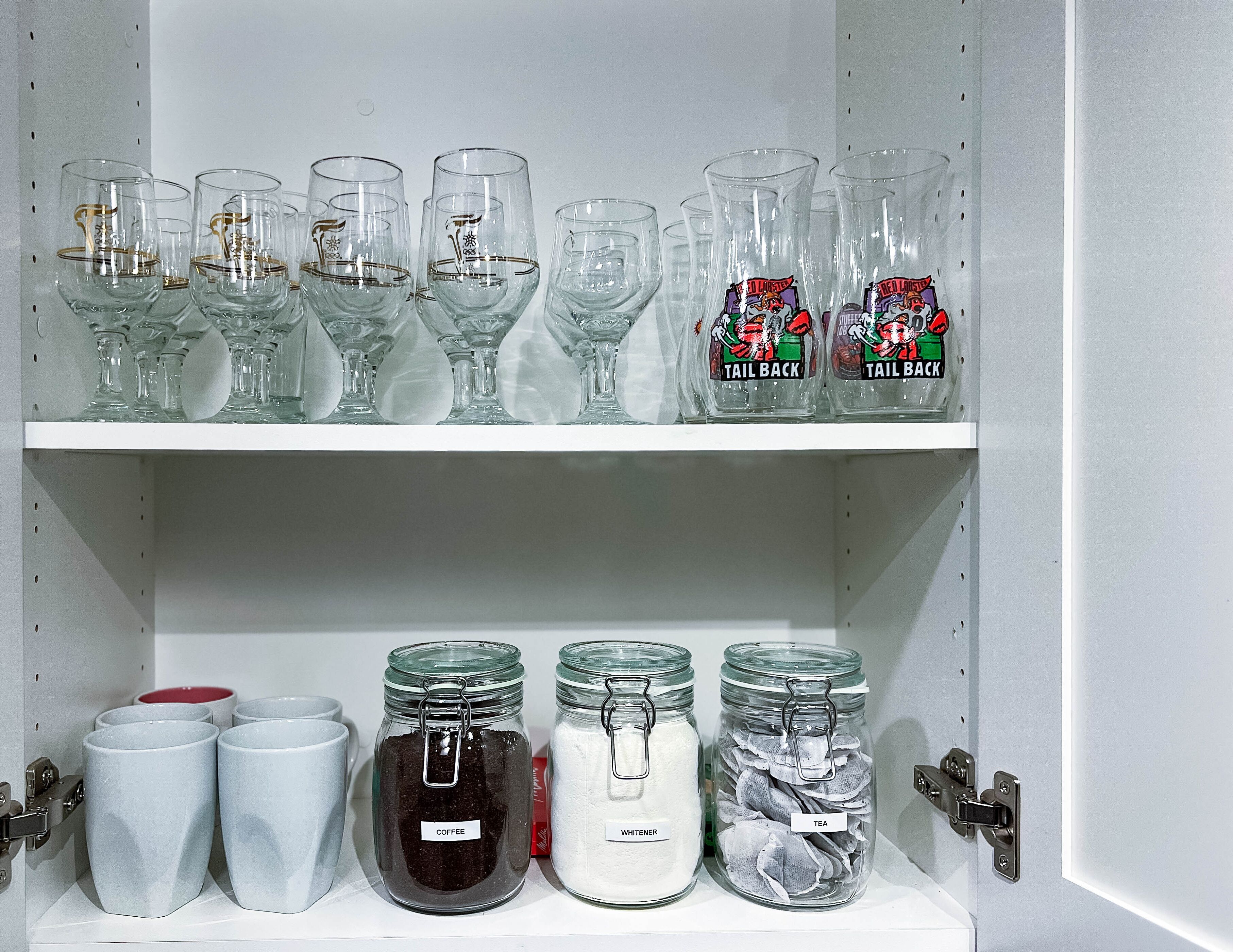 En hylde med forskelligt glas og glasbeholdere mærket med indhold som kaffe, te og sukker.