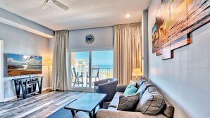 A modern living room with a sofa, armchairs, a coffee table, and a TV. There is a balcony with a view of the sea and buildings. - Madeira Bay Resort I 1502 (Madeira Beach)