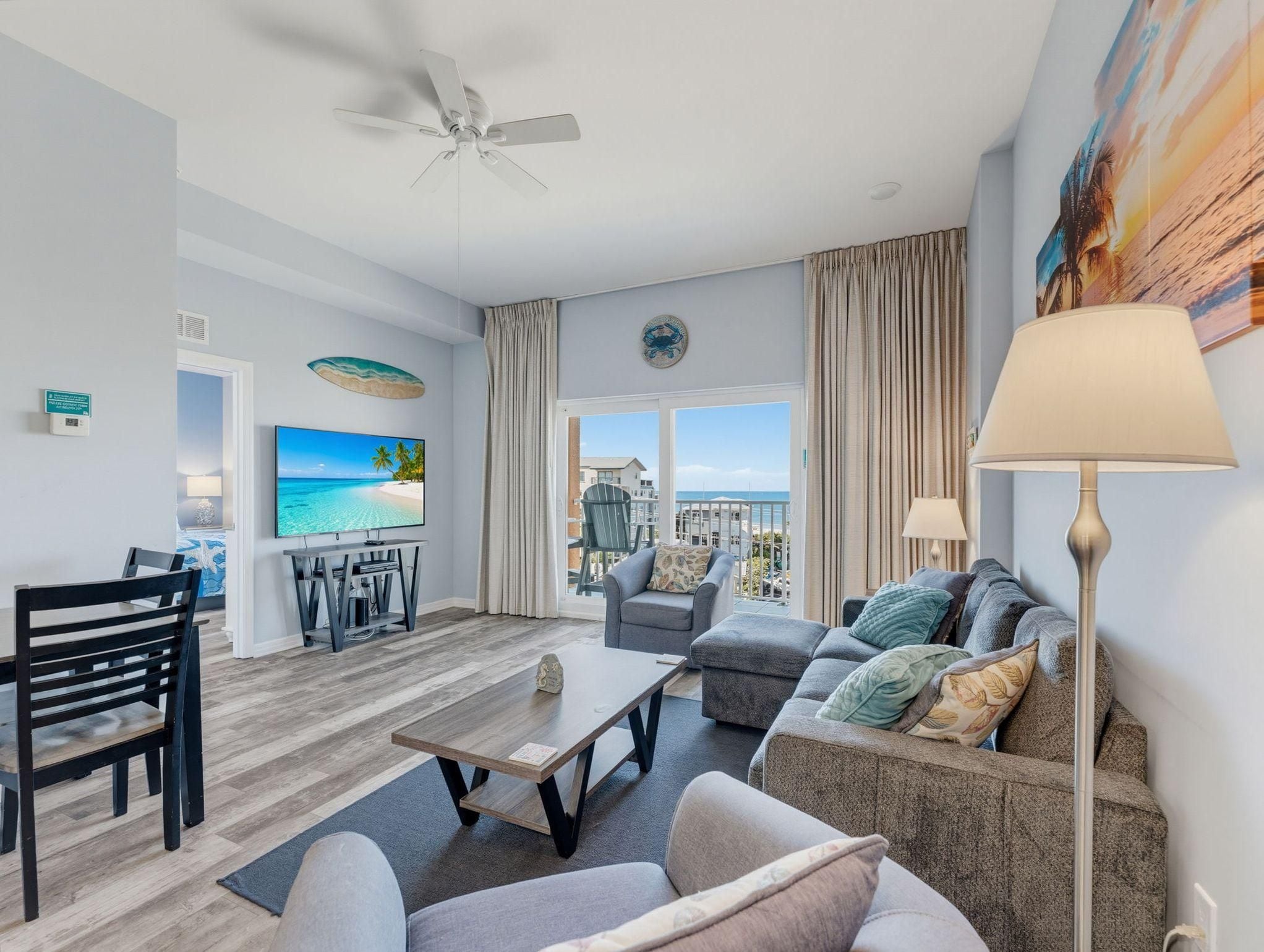 A modern living room with a flat-screen TV, a ceiling fan, a dining area with a table and chairs, and a view of the ocean through large windows.
