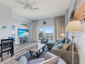 Un salon moderne doté d’un téléviseur à écran plat, d’un ventilateur de plafond, d’un coin repas avec une table et des chaises, et offrant une vue sur l’océan grâce à de grandes fenêtres.