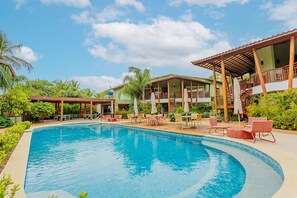 Ein Pool mit Liegestühlen und Sonnenschirmen, umgeben von Grün und einem Gebäude mit Balkon.
