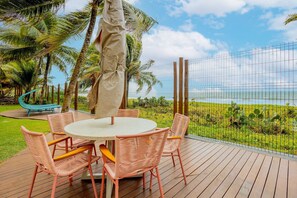 Eine Holzterrasse mit einem runden Tisch und rosa Korbstühlen, mit Blick auf einen Strand mit Palmen und einem Zaun.