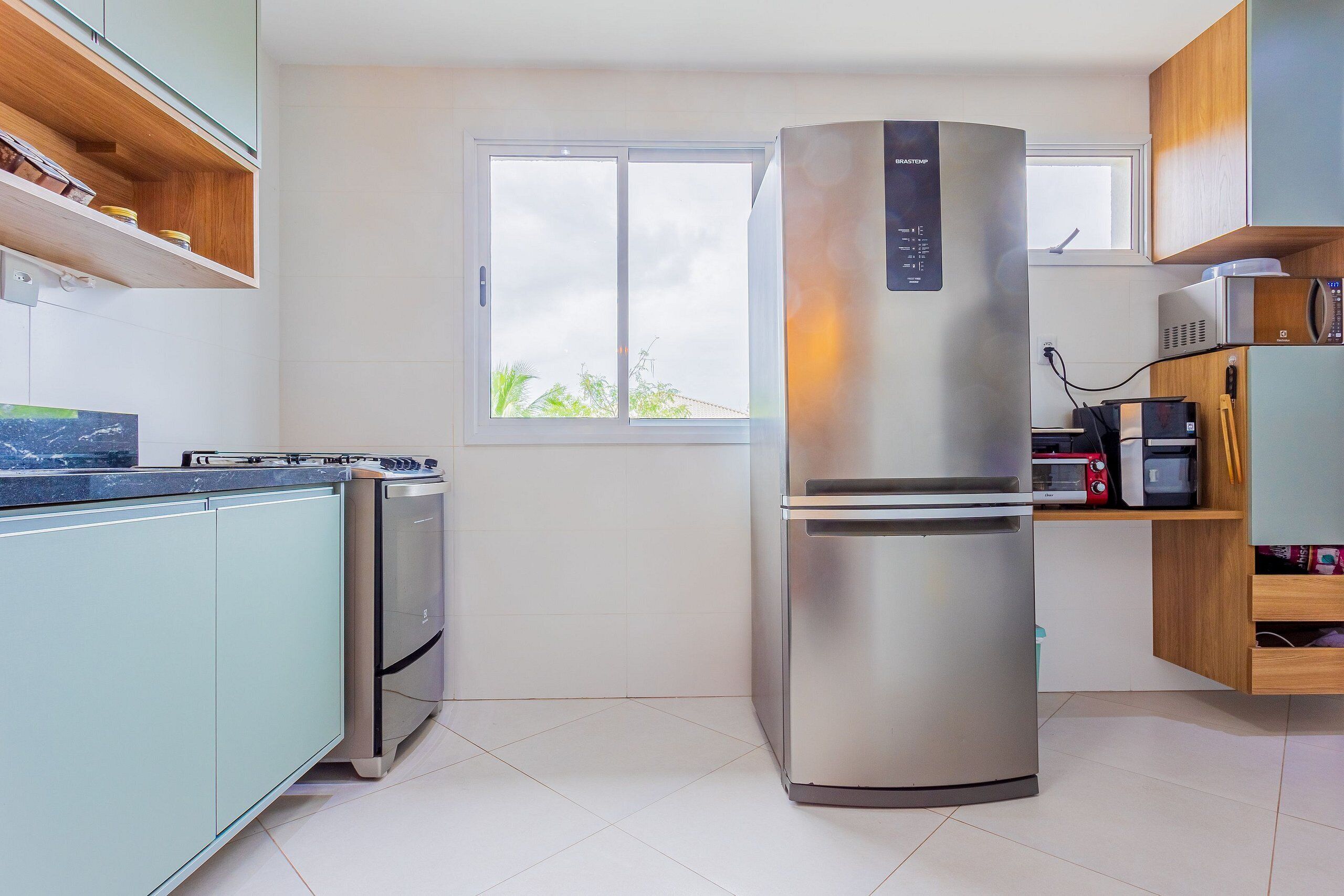 Una cocina moderna con un refrigerador de acero inoxidable, un horno empotrado y un microondas. Hay una ventana que proporciona luz natural, y repisas de madera sobre la encimera.