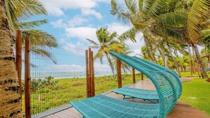 A curved blue lounge chair on a wooden deck with a view of a fenced area and palm trees. - IT22 Lindo Apto 3 Quartos a Beira Mar (Camaçari)