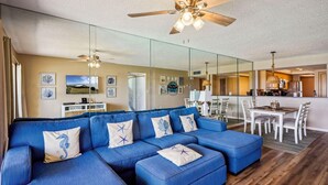 Un salon moderne avec un canapé d’angle bleu, un ventilateur de plafond et une cuisine en arrière-plan.