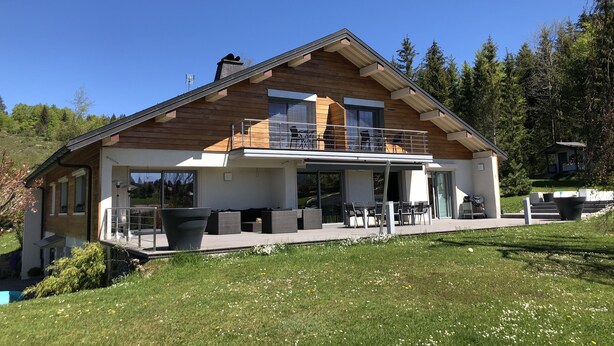 A modern two-story house with a balcony, large windows, and a patio area with outdoor furniture.