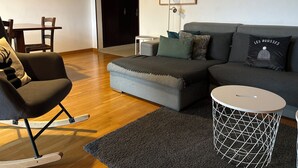 A modern living room with a grey sofa, a rocking chair, a small round table, and a framed picture on the wall.