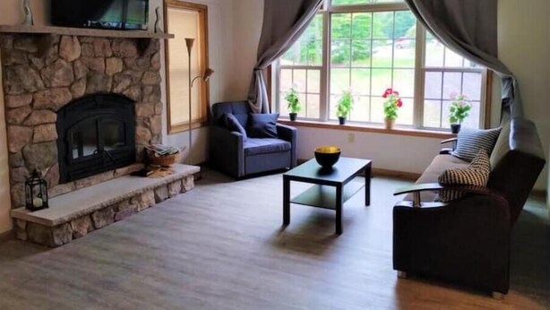 A living room with a stone fireplace, a television mounted above it, a sofa, a coffee table, and a window with curtains. - Spacious 3-bedroom house in the Poconos. (Blakeslee)
