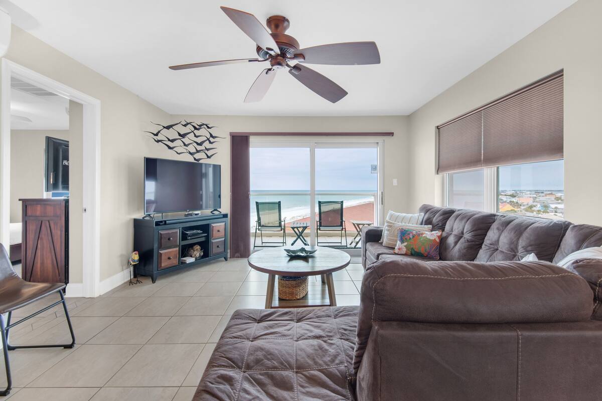 A living room with a large brown sofa, a television, a ceiling fan, and a view of the ocean through a sliding glass door.