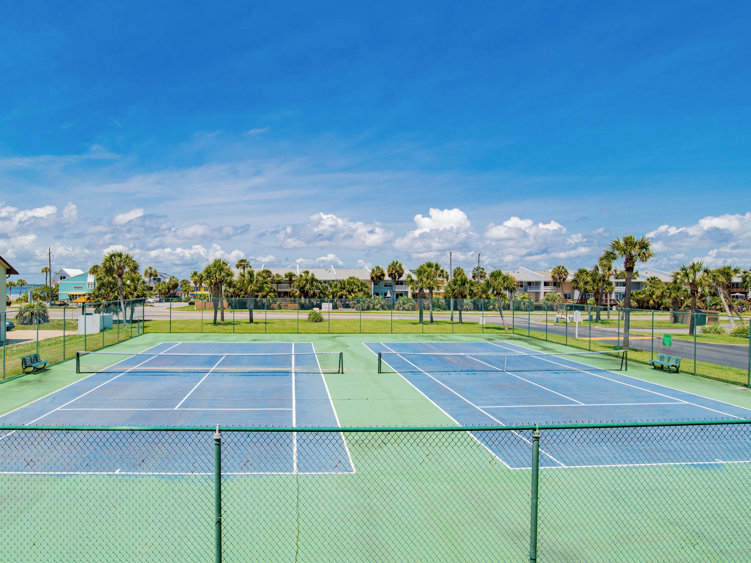 Een tennisbaan met twee velden, omringd door een hek, en palmbomen op de achtergrond.
