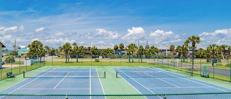 Een tennisbaan met twee velden, omringd door een hek, en palmbomen op de achtergrond.