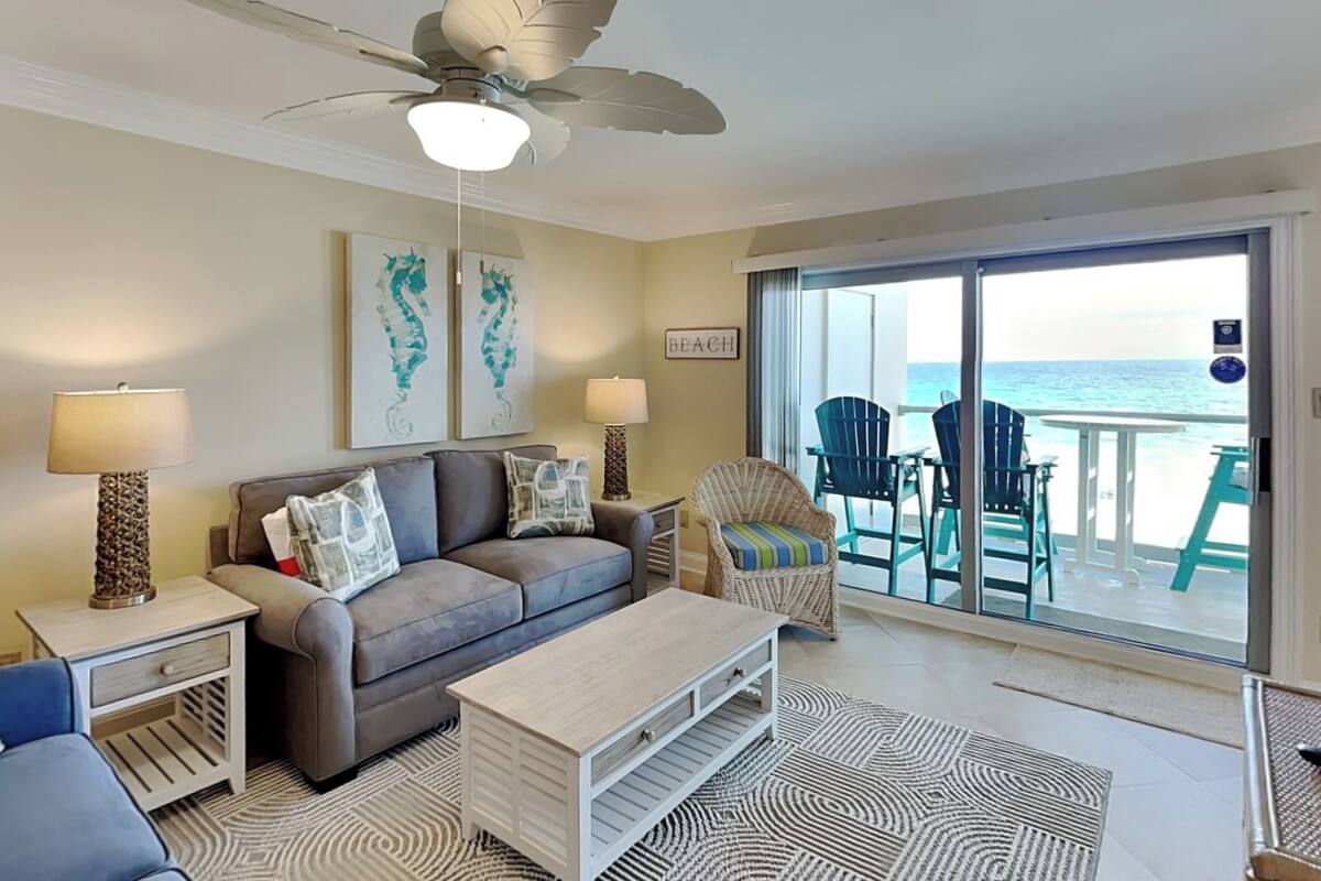 A living room with a sofa, coffee table, and chairs, a ceiling fan, and a view of the beach through a sliding glass door.