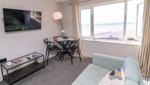 A modern living room with a sofa, a TV, a dining table with chairs, a coffee table, and a large window with a beach view. - Apt 8 @ Hunter's Quay (Bridlington)