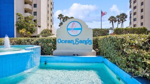 A sign for Ocean Sands hotel with a pool area, surrounded by tall buildings and palm trees.