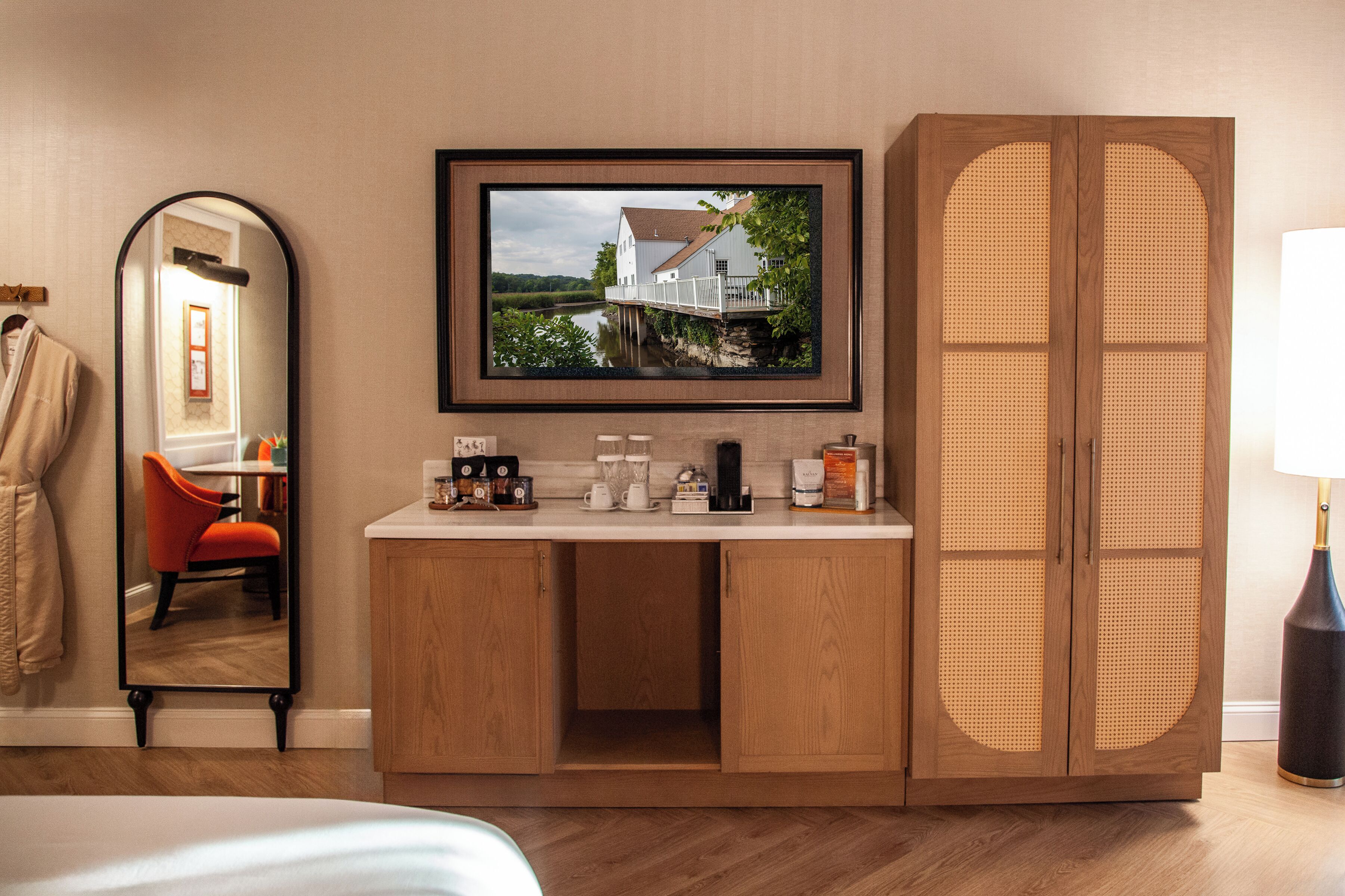 A hotel room with a wooden cabinet, a framed picture of a house, a mirror, a red chair, and a bed.