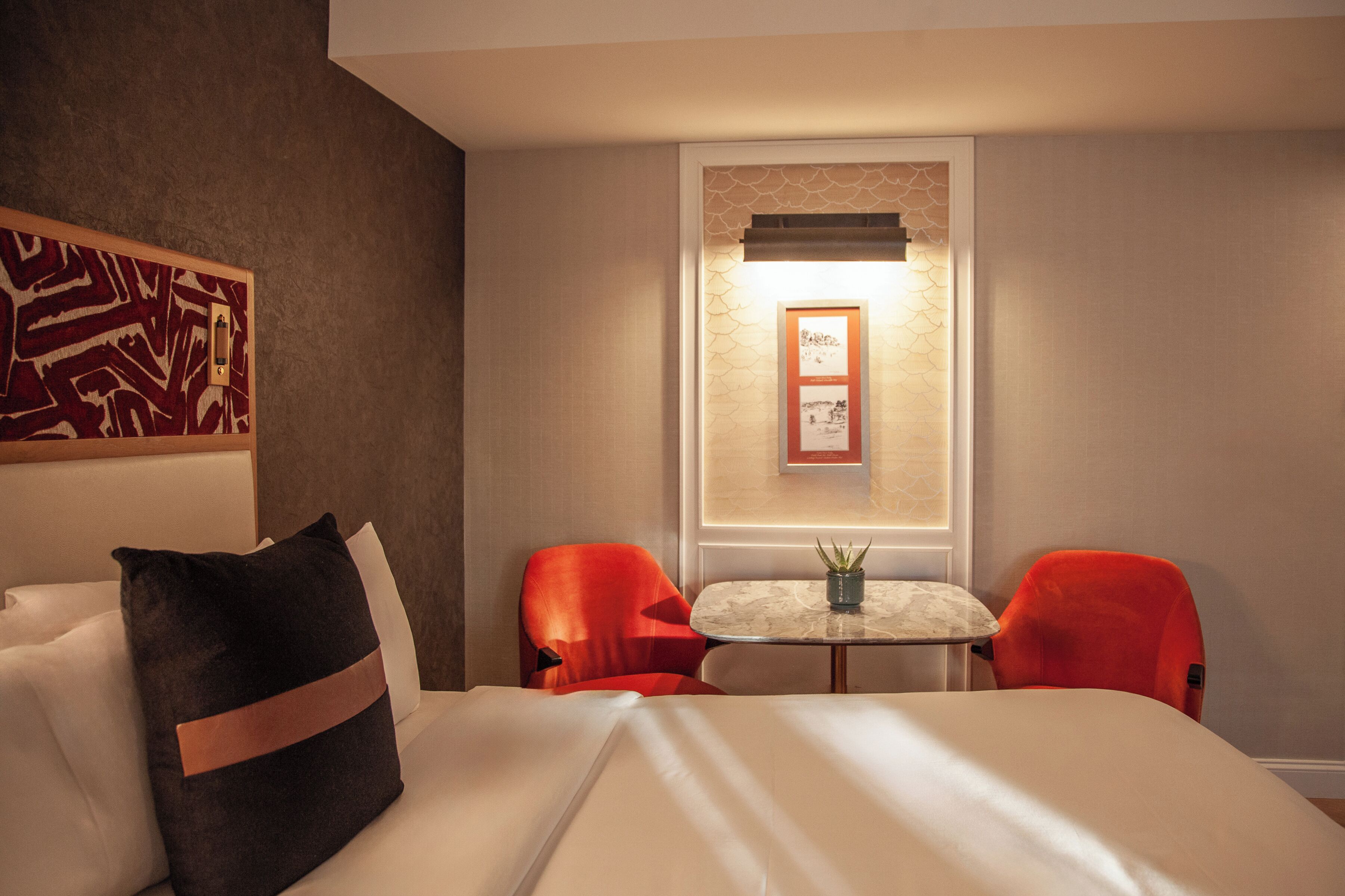 A hotel room with a bed, two red chairs, a small table with a plant, and a framed picture on the wall.