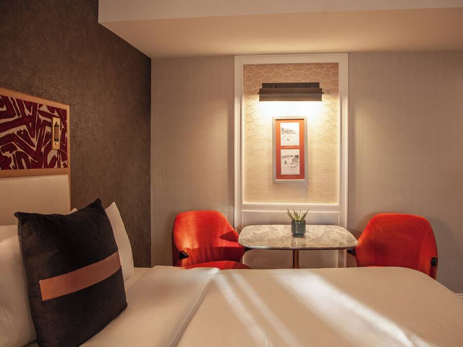 A hotel room with a bed, two red chairs, a small table with a plant, and a framed picture on the wall.