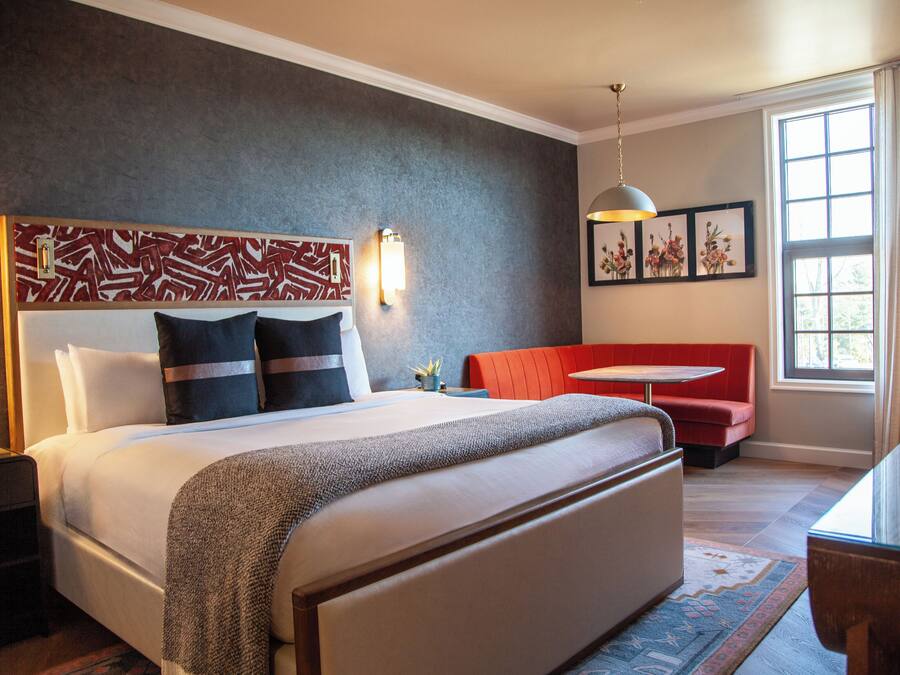 Una habitación de hotel moderna con una cama grande, un banco rojo, una mesita y un amplio ventanal con cortinas.