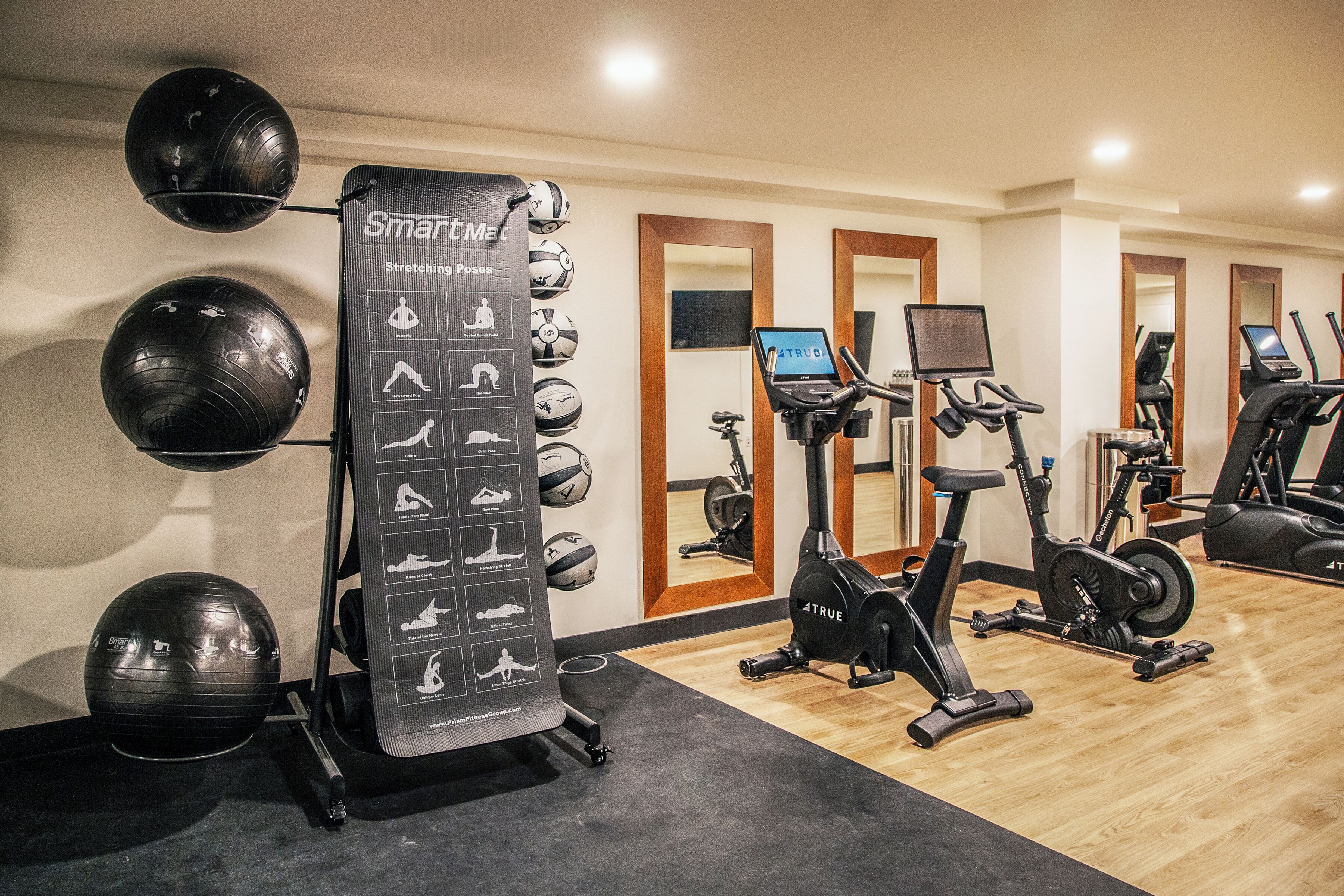 Uma academia com equipamentos de ginástica, incluindo uma máquina de remo, um simulador elíptico e um suporte para bola de exercícios fixado na parede.