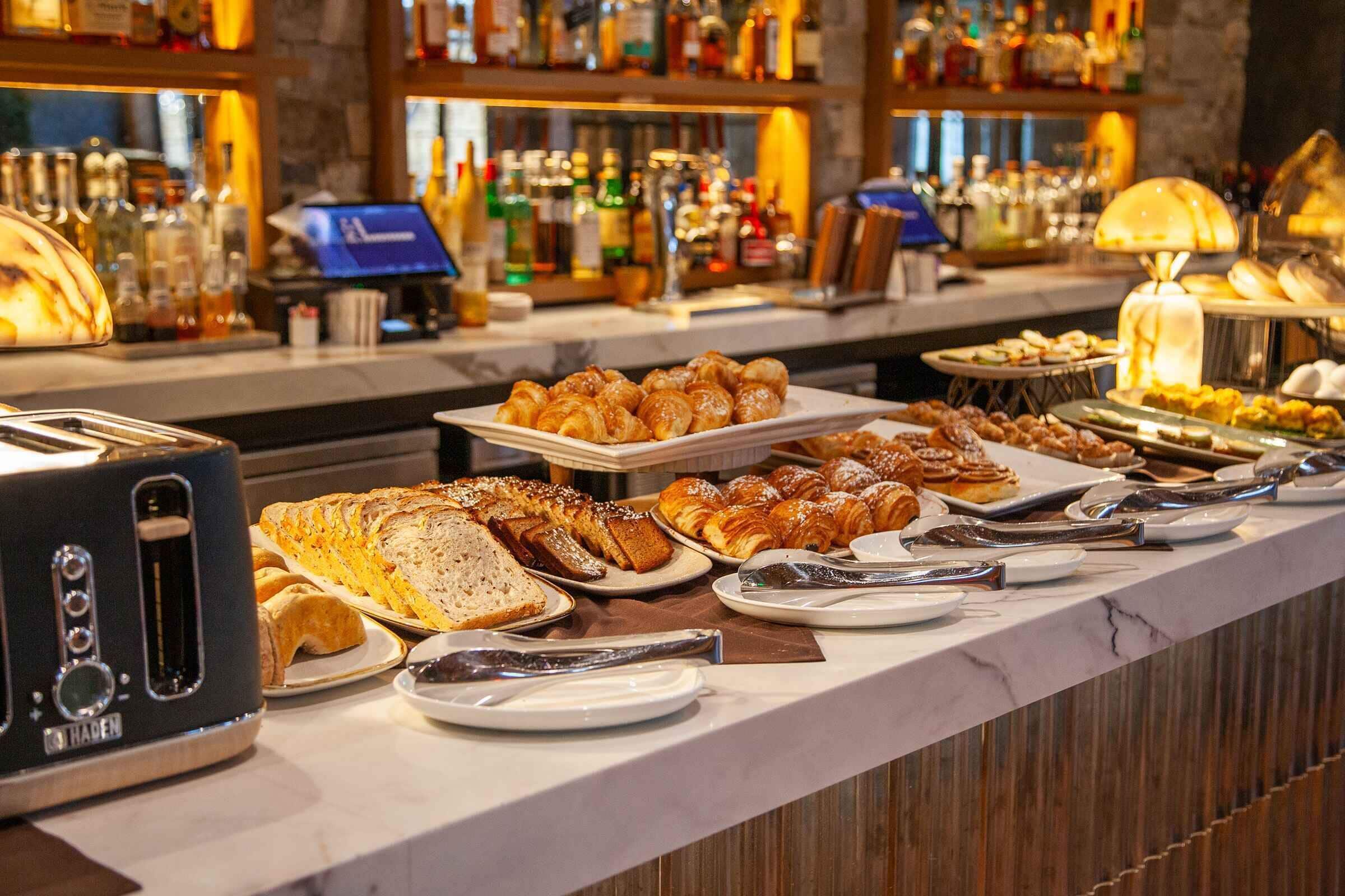 Een buffet met diverse gebakjes en brood, een kassa, en een bar met flessen op de achtergrond.