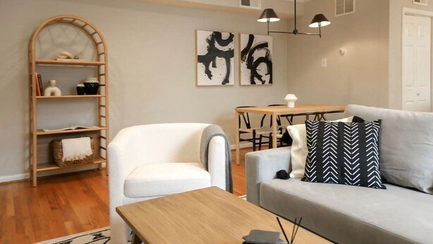 Una sala de estar moderna con un sofá blanco, una mesa de centro de madera y una estantería. Hay cuadros abstractos en la pared y una lámpara sobre la mesa.