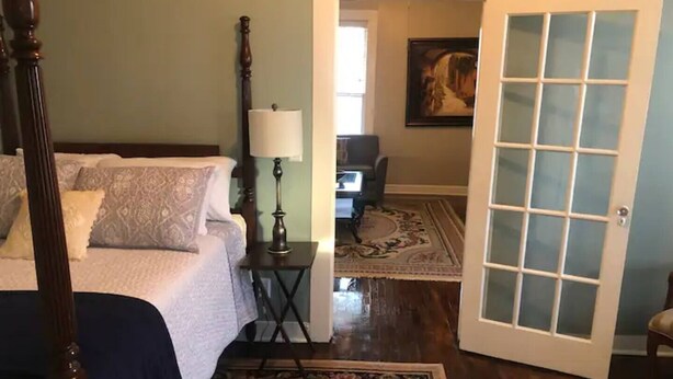 A bedroom with a four-poster bed, a bedside table with a lamp, and a chair in the corner. - The Alexandria House #1 (Jackson)
