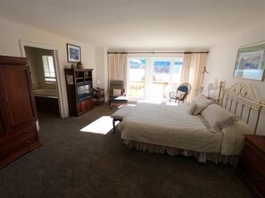 A bedroom with a large bed, a wooden wardrobe, a desk, and a chair. There is a balcony with a view.