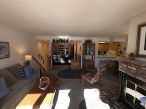 A living room with a sofa, a coffee table, and a fireplace. There is a kitchen in the background with a counter and cabinets.