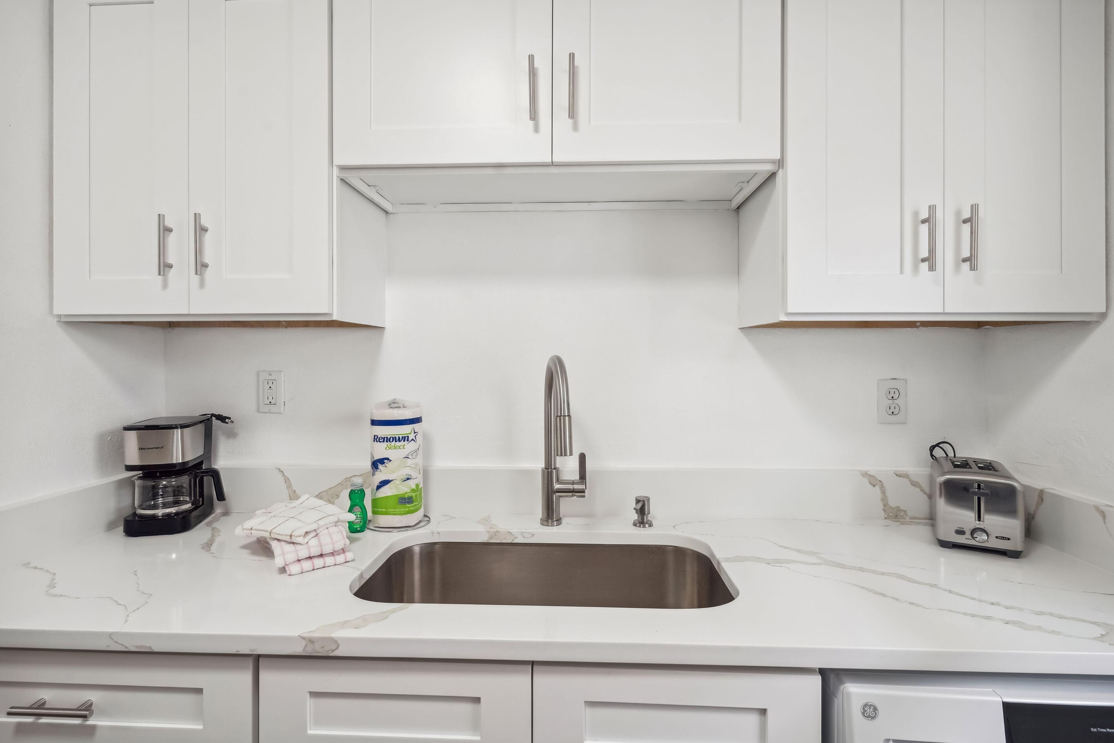 Una cocina con armarios blancos, un lavamanos de acero inoxidable, una cafetera, una botella de detergente y un tostador.