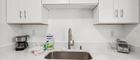 Una cocina con armarios blancos, un lavamanos de acero inoxidable, una cafetera, una botella de detergente y un tostador.