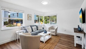 A modern living room with a grey sofa, white coffee table, and a view of a blue house through large windows. - Beach Livin 1 - 1 Bedroom Downstairs (Treasure Island)