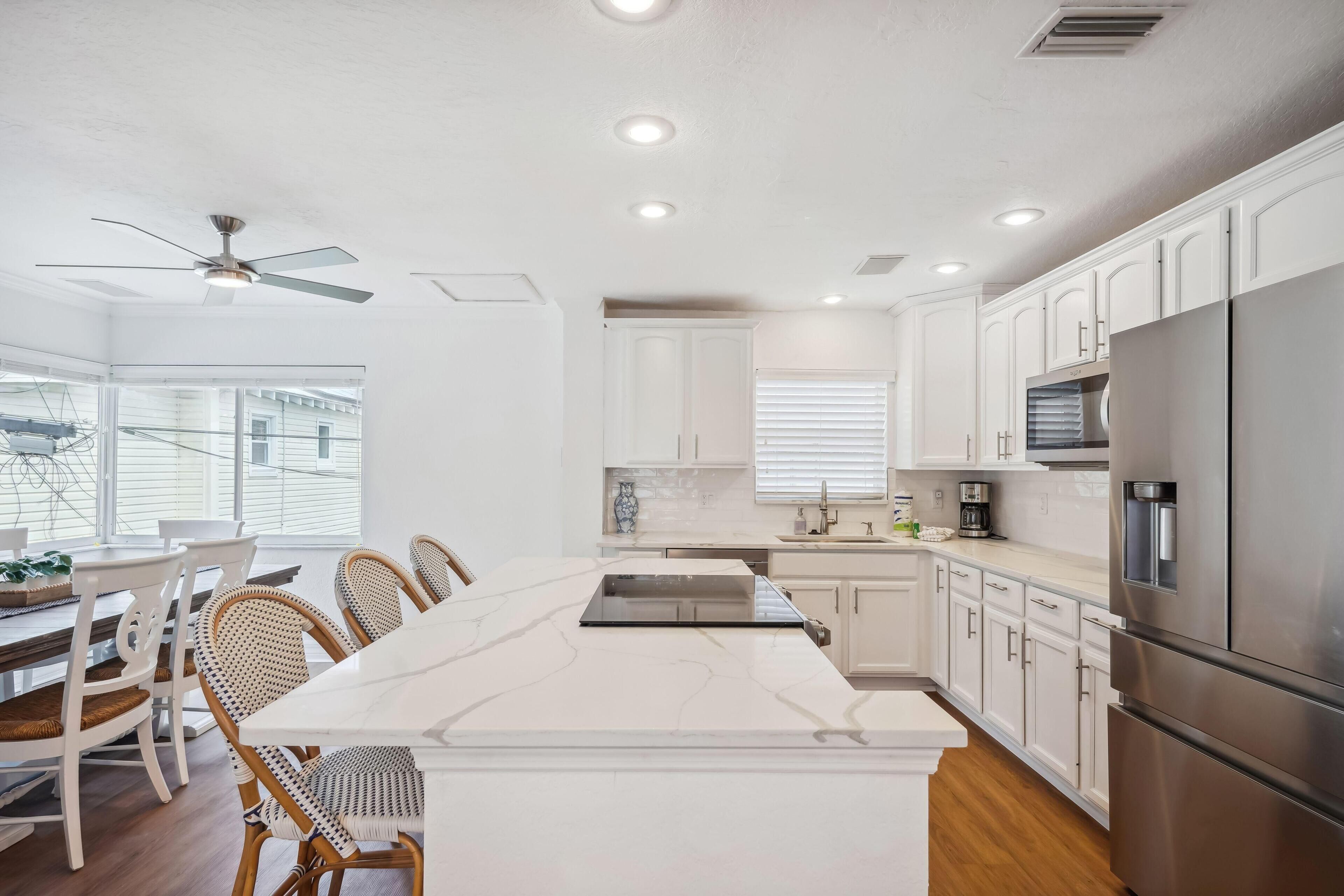 A modern kitchen with white cabinets, a stainless steel refrigerator, and a dining area with wicker chairs and a white marble island.