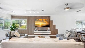 A modern living room with a large flat-screen TV mounted on a wooden panel wall, a sofa, and a dining area with a table and chairs. - Beach Livin 3 - 3 Bedroom Upstairs (Treasure Island)