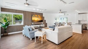 A modern living room with a large TV, a white sofa, a blue sofa, and a dining area with a table and chairs. - Beach Livin - All (Treasure Island)