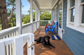A wooden deck with a dog lying on the floor, a porch with white railings, and colorful chairs. - Spacious, Dogs Welcome, 5-mins to Beach (Waves)