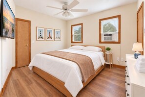 A bedroom with a bed, a ceiling fan, framed pictures, a window, and a bedside table with a lamp. - Spacious, Dogs Welcome, 5-mins to Beach (Waves)