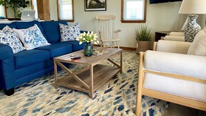 A living room with a blue sofa, a wooden coffee table, a white armchair, and a beige sofa. There is a TV mounted on the wall, a plant, and a framed picture on the wall.