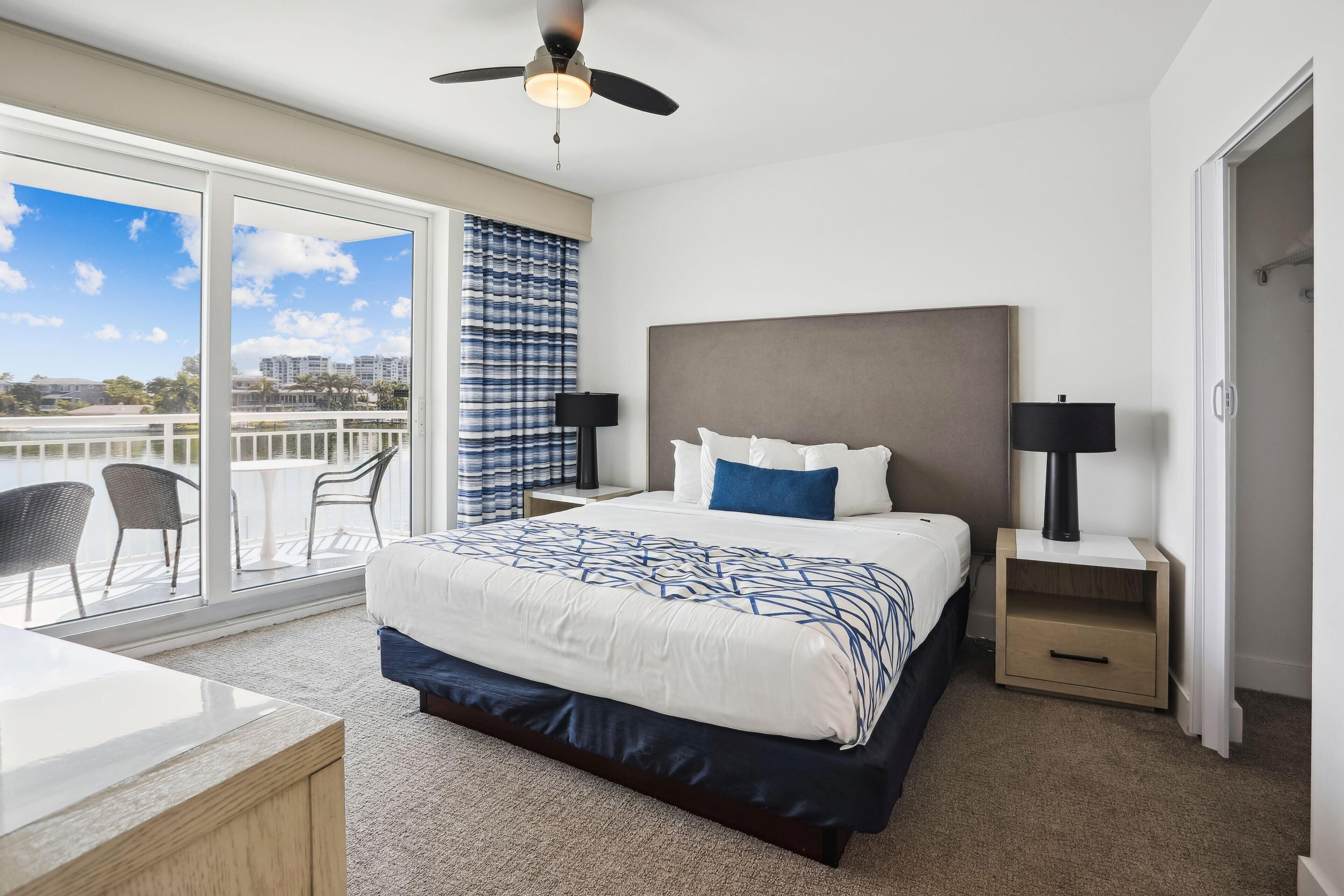 Una habitación de hotel moderna con una cama grande, ventilador de techo, mesita de noche, escritorio y vistas a la ciudad a través de amplias ventanas.