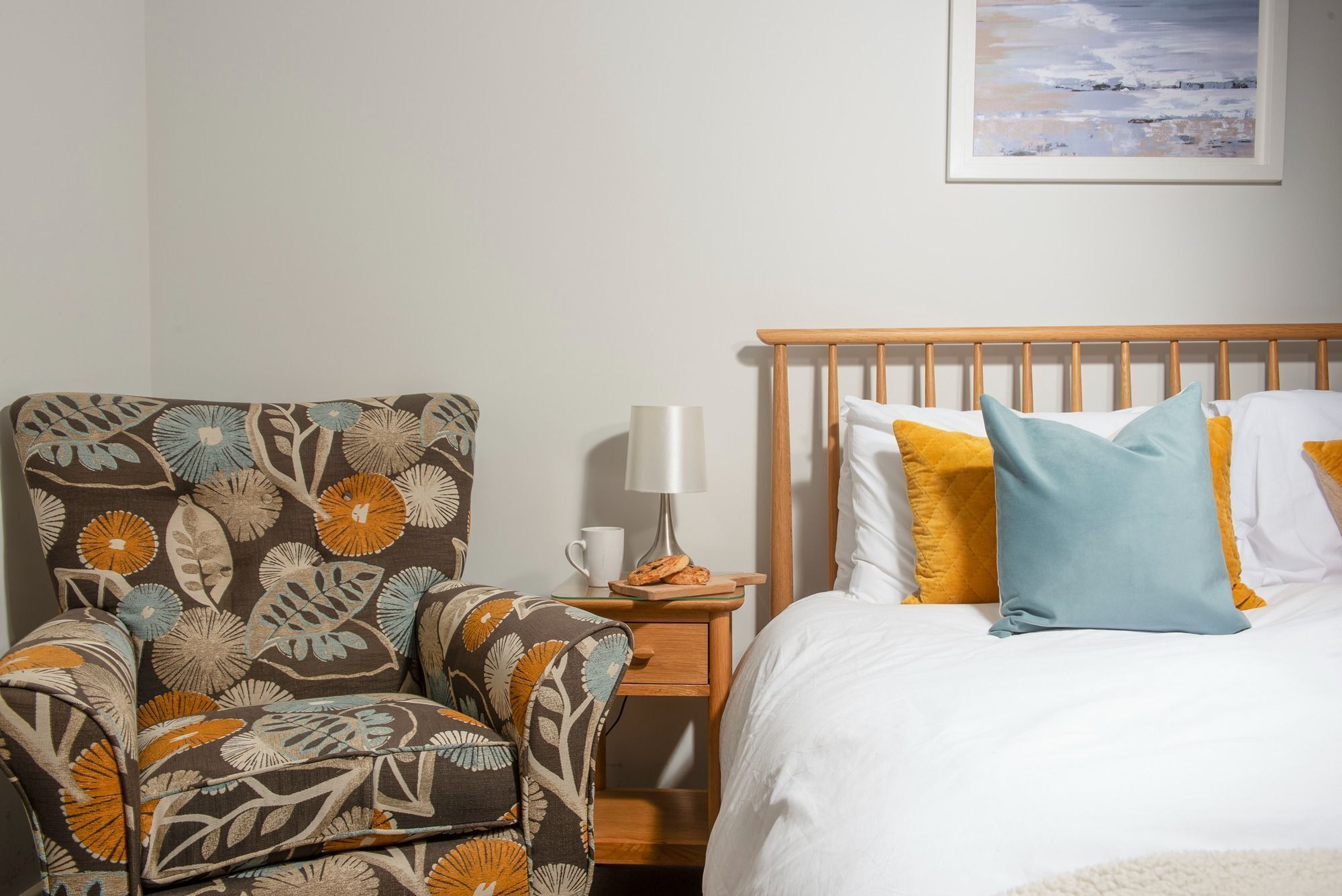 Een slaapkamer met een houten bed, een gestreept fauteuil, een nachtkastje met een lamp en een lijst met een afbeelding aan de muur.