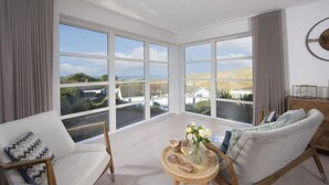 Een ruime kamer met grote ramen en uitzicht op het landschap. Er staat een bank, een stoel en een klein houten tafeltje met een bloevas.